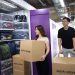 A man and woman move cardboard boxes into a storage unit with bright purple doors, filled with neatly organized plastic bins and gear